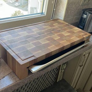 Puede incluir: Una tabla de cortar rectangular de madera de testa con un patrón de tablero de ajedrez de bloques de madera de color marrón claro y medio. La tabla tiene un perfil grueso con una ranura empotrada en el borde superior y una bandeja de metal.