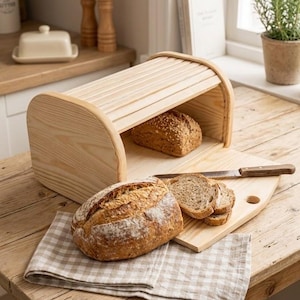 Puede incluir: Una panera de madera con tapa enrollable, que contiene un pan. Un pan y pan en rodajas están sobre una tabla de cortar de madera con un cuchillo. Un paño a cuadros está debajo del pan. Escena de cocina rústica.