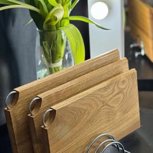 Puede incluir: Un conjunto de tres tablas de cortar rectangulares de madera con un acabado de vetas naturales. Cada tabla tiene un asa de acero inoxidable y se exhibe en un estante de alambre cromado. Un jarrón con hojas verdes y un tulipán amarillo en el fondo.
