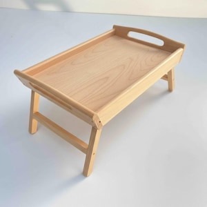 May include: A light-colored wooden breakfast tray with folding legs. The tray has a rectangular surface with a raised edge and cut-out handles. The legs are angled for stability.