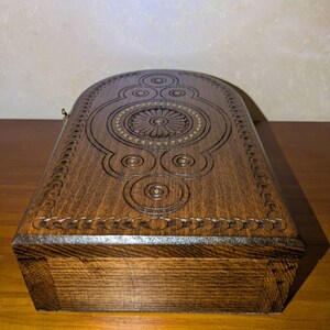 May include: A dark brown wooden box with a rounded top edge. The lid features intricate carved geometric patterns, including a central circular floral motif surrounded by smaller concentric circles and a decorative border along the curved edge.