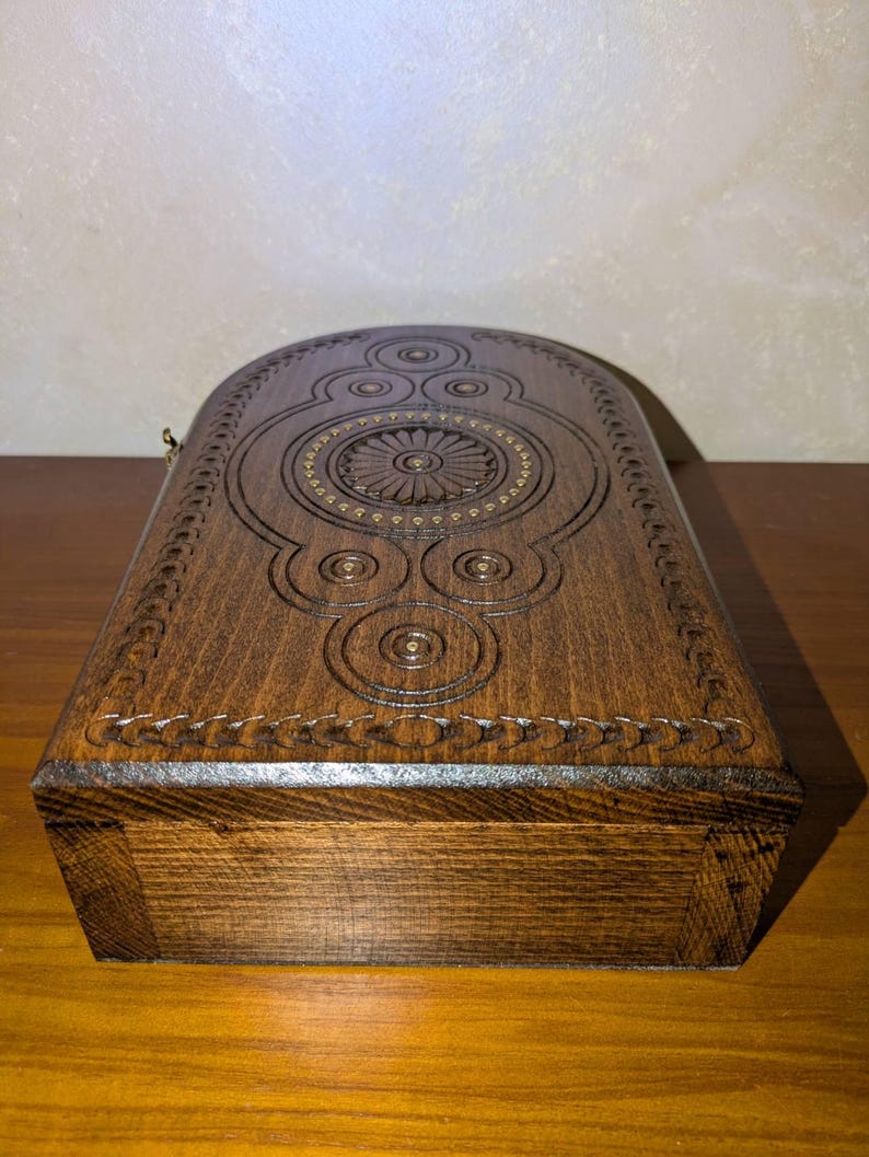 May include: A dark brown wooden box with a rounded top edge. The lid features intricate carved geometric patterns, including a central circular floral motif surrounded by smaller concentric circles and a decorative border along the curved edge.