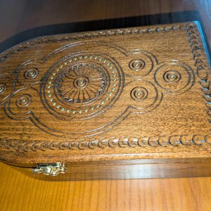 May include: A rectangular wooden box with a rounded end, featuring intricate carved geometric patterns and a central circular motif accented with small brass studs. The box has a polished brown finish and a metallic latch on the front edge.