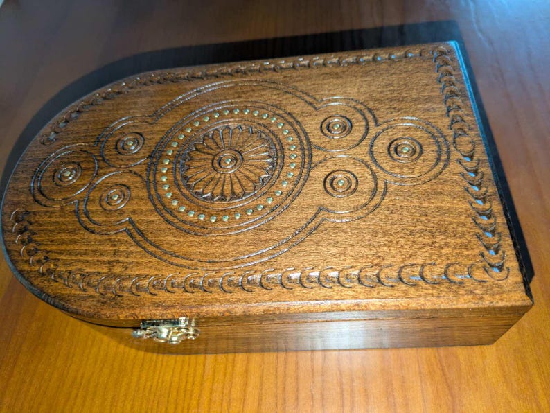 May include: A rectangular wooden box with a rounded end, featuring intricate carved geometric patterns and a central circular motif accented with small brass studs. The box has a polished brown finish and a metallic latch on the front edge.
