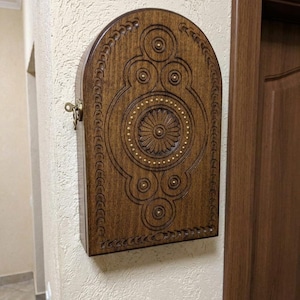 May include: A dark brown wooden wall-mounted cabinet with an arched top. The front panel features intricate carved geometric patterns, circular motifs, and a central flower design accented with small brass studs. A brass key is inserted in the side.