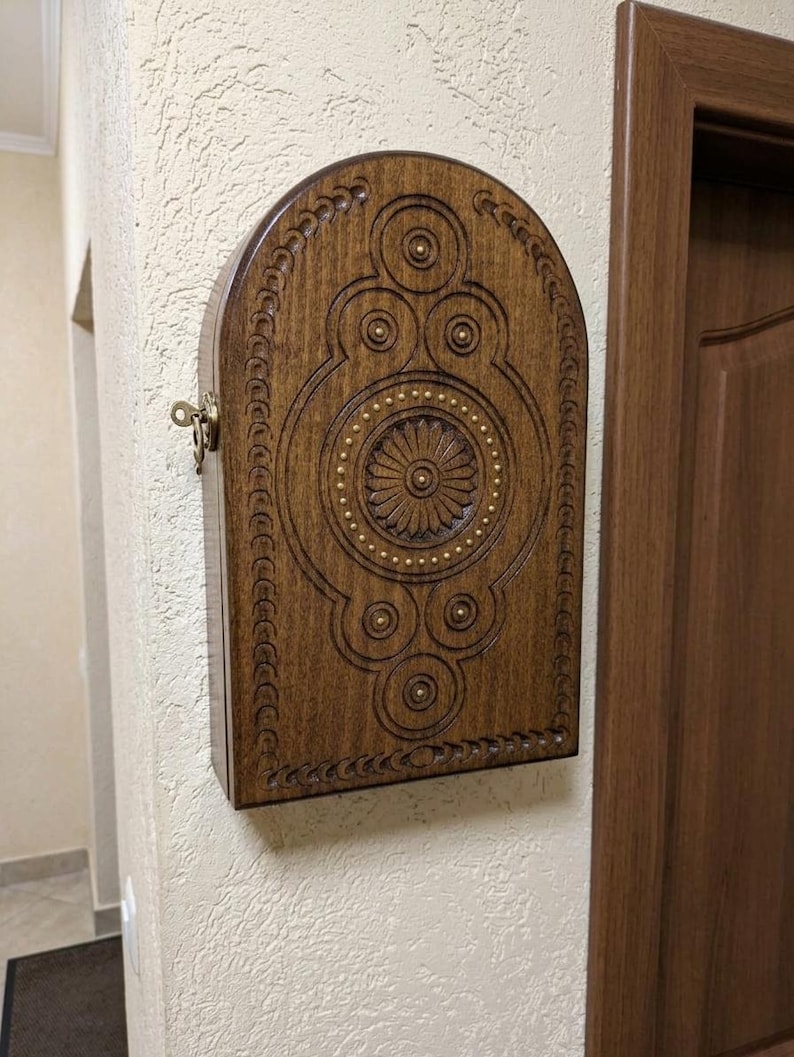 May include: A dark brown wooden wall-mounted cabinet with an arched top. The front panel features intricate carved geometric patterns, circular motifs, and a central flower design accented with small brass studs. A brass key is inserted in the side.