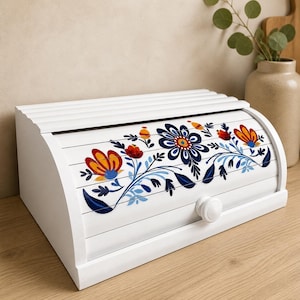 May include: White wooden bread box with a roll-top lid. The front is decorated with a floral design in blue, red, and yellow. The box has a white knob and is on a wooden surface.