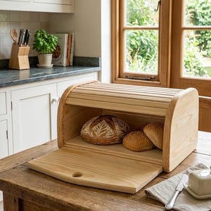 Peut inclure: Un coffre à pain en bois clair avec un couvercle roulant, ouvert pour révéler deux pains. Le coffre est posé sur une table en bois. Un beurrier et un couteau sont également sur la table.