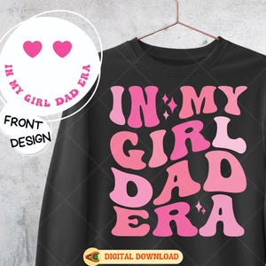 May include: Black sweatshirt with the phrase "IN MY GIRL DAD ERA" in shades of pink. The design includes stars and a smiley face with two pink hearts. The words "FRONT DESIGN" are also visible.