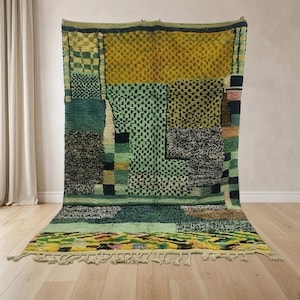 Könnte beinhalten: Ein großer, rechteckiger Teppich mit einem Patchwork-Design. Der Teppich weist eine Vielzahl von Farben auf, darunter Grüntöne, Gelb und Schwarz. Das Design umfasst geometrische Muster und einen Fransenbesatz am unteren Rand. Der Teppich wird auf einem Holzboden präsentiert.