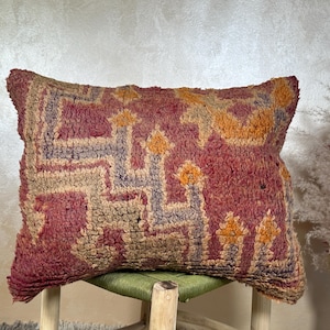 Op de afbeelding: Een rechthoekig, getextureerd kussen met een opvallend geometrisch patroon. Het kussen heeft een dieprode basiskleur met accenten van oranje, beige en blauw. Het ligt op een groene, geweven zitting.