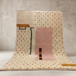 May include: A rectangular area rug featuring a cream-colored base with a repeating pattern of small gold diamond shapes. The design includes a large central pink block, a horizontal gold bar, a vertical black line, and a small dark brown striped accent.