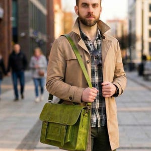 Op de afbeelding: Een felgroene leren tas met een voorflap, twee verticale gespriemen en een bijpassende verstelbare schouderriem. De tas is voorzien van een rechthoekig voorvak en metalen hardware en wordt over een beige trenchcoat gedragen.