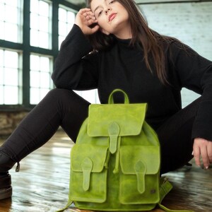 May include: A bright green leather backpack with two front pockets and a flap closure. The backpack has a top handle and adjustable straps. The bag is sitting on a wooden floor.