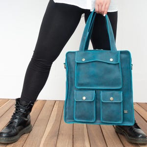 Op de afbeelding: Een teal leren tote bag met meerdere vakken en een handvat aan de bovenkant. De tas heeft een rechthoekige vorm met een flap en een knoopsluiting op de voorzakken. De tas is afgewerkt met teal stiksels.