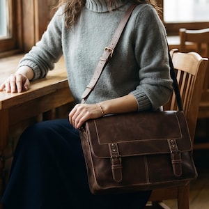 Puede incluir: Un bolso tipo mensajero de cuero marrón oscuro con una solapa rectangular, dos correas frontales verticales con hebillas metálicas y una correa ajustable para el hombro. El bolso presenta costuras visibles en los bordes y un acabado mate.
