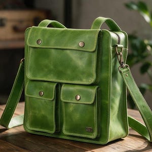 Op de afbeelding: Een vierkante groene leren tas met een grote klepzak aan de bovenkant en twee kleinere zakken aan de onderkant, elk beveiligd met metalen drukknopen. De tas is voorzien van een afneembare schouderriem met metalen hardware en stiksels.