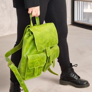 Puede incluir: Una mochila de cuero verde lima con dos bolsillos delanteros y correas ajustables. La mochila es sostenida por una persona que lleva pantalones negros y botas negras.