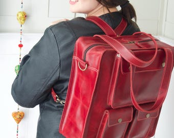 May include: A red leather backpack with multiple pockets and white stitching. The bag has a top handle and adjustable straps. The person in the image is wearing a black hat and jacket. The bag is a vibrant red color.