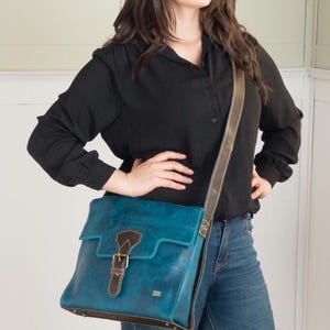 May include: A teal leather messenger bag with a brown leather trim and strap. The bag has a buckle closure and a flap. The bag is worn over the shoulder. The bag is a satchel style.