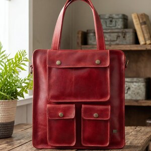 Peut inclure: Un sac fourre-tout en cuir rouge profond doté de deux anses supérieures et de trois poches extérieures avec des fermetures à pression couleur laiton. Le sac a une forme rectangulaire avec des coutures visibles et une petite étiquette métallique.