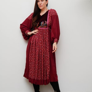Może przedstawiać: Bordowa sukienka midi z długimi rękawami i aksamitnym stanikiem. Sukienka posiada haftowane kwiaty i spódnicę w kropki. Modelka ma na sobie czarne buty.