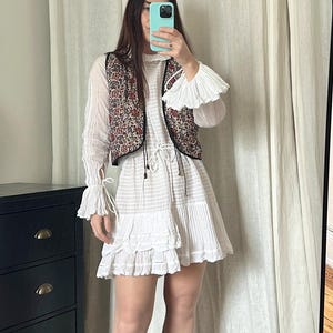 Puede incluir: Un vestido blanco con volantes en capas, mangas largas con puños con volantes y un chaleco floral. El atuendo se combina con botines marrones. La imagen se toma frente a un espejo.