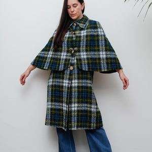 May include: A knee-length cape coat featuring a green, blue, and white plaid pattern. The garment has a fold-over collar, a button-down front with metallic leaf-shaped fasteners, and a wide, flared silhouette that extends over the shoulders.