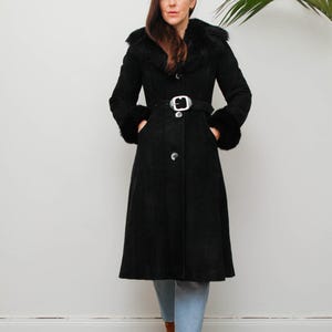 Op de afbeelding: Een lange zwarte suède jas met een kraag en manchetten van imitatiebont. De jas heeft een ceintuur in de taille met een zilveren gesp en knoopsluitingen. Het model draagt een blauwe jeans en bruine laarzen.