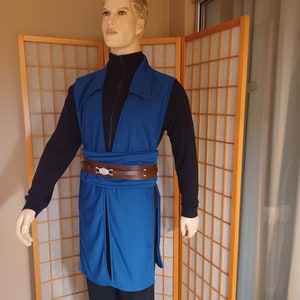 May include: A blue tunic with a brown leather belt and silver buckle. The tunic has a V-neck and long sleeves. The person is wearing black pants.
