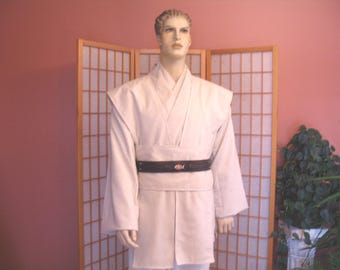 May include: A light beige Jedi-style costume displayed on a mannequin. The costume includes a long-sleeved tunic, a wrap-around belt with a decorative emblem, and matching pants. The mannequin stands in front of a folding screen and a pink wall.