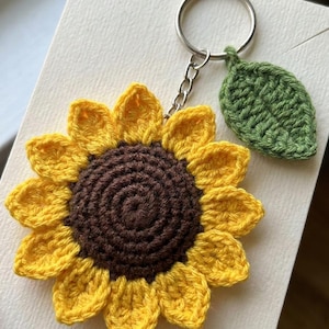 May include: A crochet sunflower keychain featuring a dark brown center and bright yellow petals. A small, textured green leaf is attached to the silver-toned metal keyring by a short chain, creating a floral accessory with distinct yarn stitching.