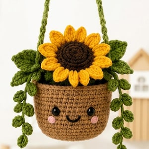 May include: A crocheted hanging plant featuring a brown pot with a smiling face, black eyes, and pink cheeks. A large yellow sunflower with a dark brown center sits in the pot, surrounded by green leaves and trailing vines hanging from the sides.