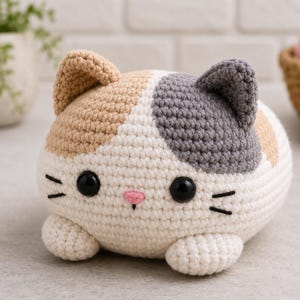 May include: A handmade crochet cat toy in cream, tan, and gray. The round cat has black safety eyes, a pink nose, and black whiskers. The cat has small, rounded ears and small paws. The toy is sitting on a light gray surface.