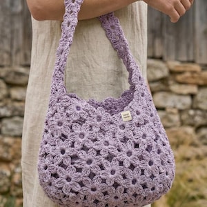 Könnte beinhalten: Eine lavendelfarbene Häkeltasche mit Blumenmuster. Die Tasche hat eine runde Form und einen passenden Träger. Ein kleines Etikett mit der Aufschrift "hand made" ist an der Tasche befestigt.