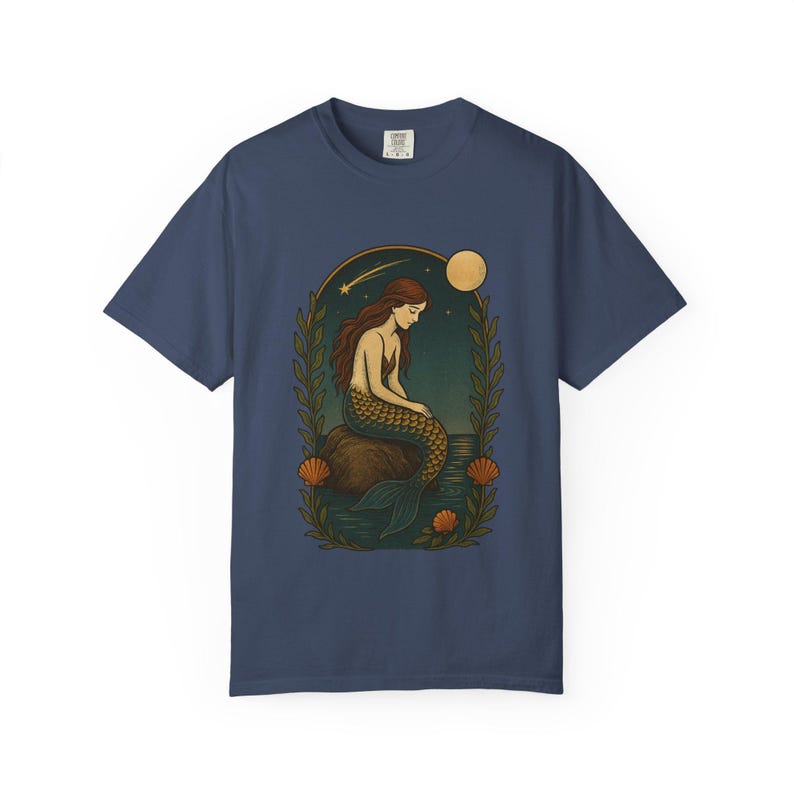 May include: Navy blue t-shirt featuring a mermaid illustration. The design shows a mermaid with long red hair and a gold and green tail, sitting on a rock. The background includes a moon, stars, and foliage. The shirt is a comfortable, casual style.