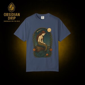 May include: A blue t-shirt featuring a mermaid illustration. The mermaid has long brown hair and a scaled tail, sitting on a rock with a moon and stars in the background. The shirt also has the text "Obsidian Drip" and "Darkwear for the Bold".