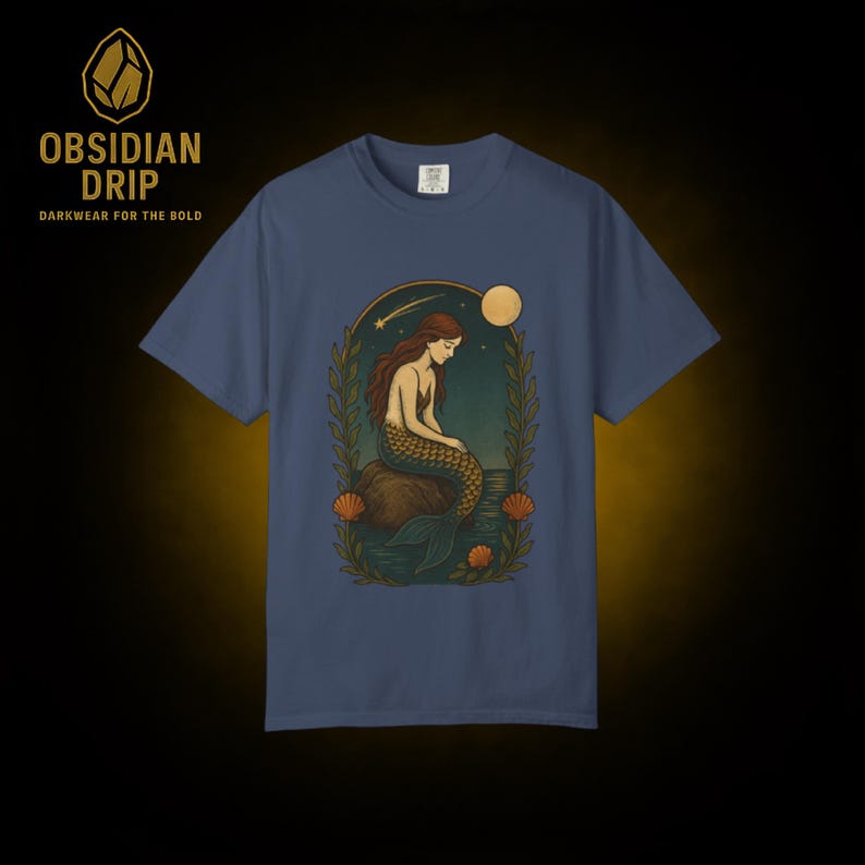 May include: A blue t-shirt featuring a mermaid illustration. The mermaid has long brown hair and a scaled tail, sitting on a rock with a moon and stars in the background. The shirt also has the text "Obsidian Drip" and "Darkwear for the Bold".