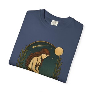 May include: Navy blue long-sleeve shirt featuring a celestial illustration of a woman with long red hair, a moon, and stars. The design is framed by green leaves and a gold-colored arch. The shirt has a crew neck and a tag.
