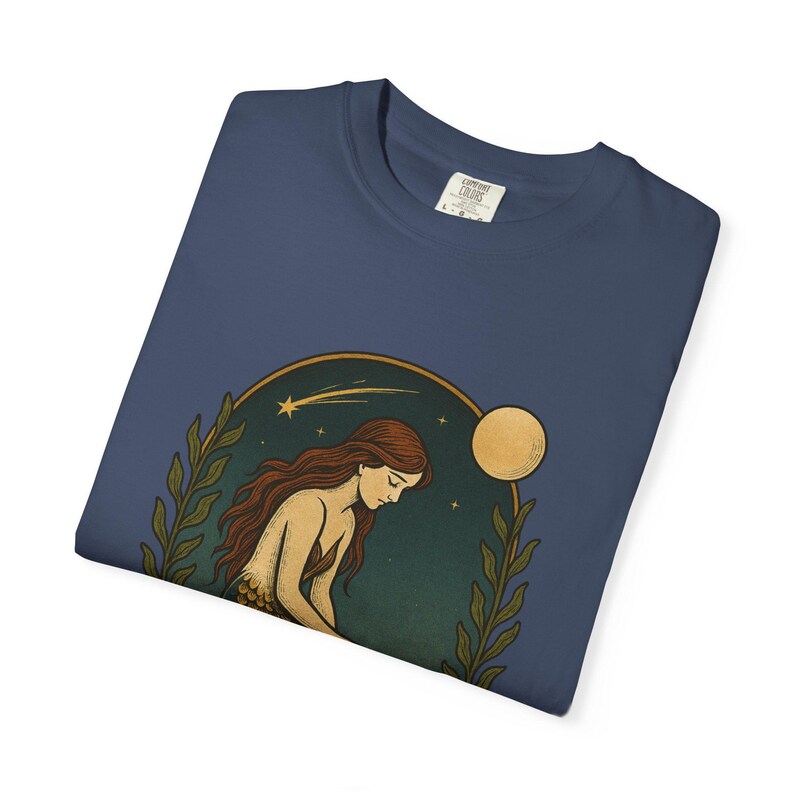 May include: Navy blue long-sleeve shirt featuring a celestial illustration of a woman with long red hair, a moon, and stars. The design is framed by green leaves and a gold-colored arch. The shirt has a crew neck and a tag.