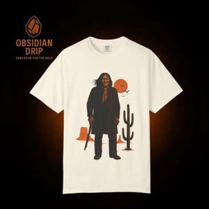May include: Cream-colored t-shirt featuring a graphic of a Native American man in a long coat, holding a staff, with a desert landscape backdrop. The shirt has the text "OBSIDIAN DRIP" and "DARKWEAR FOR THE BOLD" in dark brown.