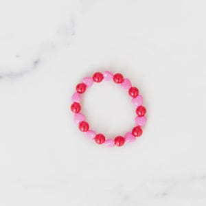 Op de afbeelding: Een armband gemaakt van roze hartvormige kralen en rode ronde kralen. De kralen wisselen elkaar af in een cirkelvormig patroon. De armband staat op een witte achtergrond. Het ontwerp van de armband is eenvoudig en elegant.