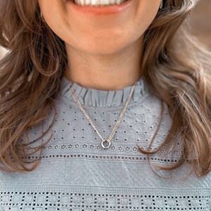 May include: A silver necklace with a delicate circle pendant, worn by a person with long brown hair. The necklace is layered over a light blue, lace-trimmed blouse.