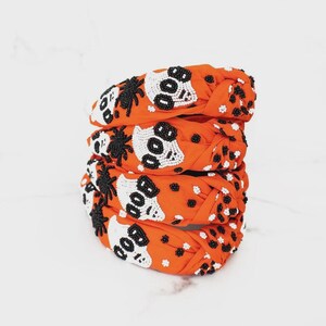 Puede incluir: Cuatro diademas naranjas de Halloween apiladas. Cada diadema presenta un diseño de calavera con cuentas con la palabra "BOO" y acentos de arañas negras. Lunares blancos salpican la tela naranja.