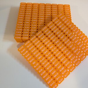 Peut inclure: Une collection de tuiles rectangulaires en plastique orange disposées en grille, chacune présentant un texte blanc imprimé composé de mots de quatre lettres commençant par A, B et C, tels que CACT, CAGE, CAKE, CALL, CALM, CAME, CAMP, CAN, AWAK, AWAR et BABY.