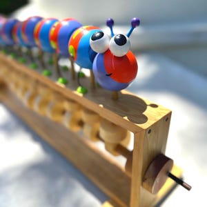 May include: A colorful wooden caterpillar toy with large googly eyes and purple antennae. The caterpillar is made of blue, red, and orange painted wooden spheres. It sits on a wooden frame with rolling cylinders, a classic educational toy.