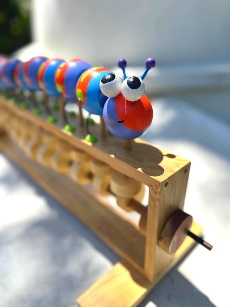 May include: A colorful wooden caterpillar toy with large googly eyes and purple antennae. The caterpillar is made of blue, red, and orange painted wooden spheres. It sits on a wooden frame with rolling cylinders, a classic educational toy.