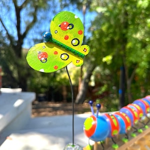 May include: A whimsical, hand-painted butterfly garden stake with a green and yellow color scheme, accented with red spots and black circles. The butterfly is mounted on a black metal rod, with a stone base. A colorful caterpillar is also visible.