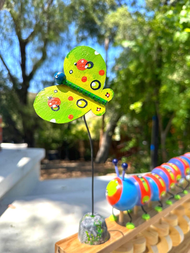 May include: A whimsical, hand-painted butterfly garden stake with a green and yellow color scheme, accented with red spots and black circles. The butterfly is mounted on a black metal rod, with a stone base. A colorful caterpillar is also visible.