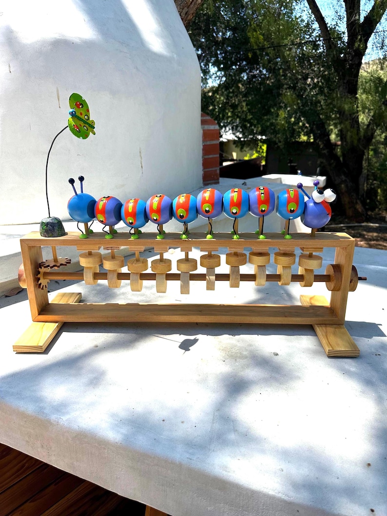 May include: A wooden abacus toy featuring a colorful caterpillar design. The caterpillar has blue, red, and yellow segments with green legs. A green and yellow butterfly is attached to the side. The abacus has wooden beads and a wooden frame.